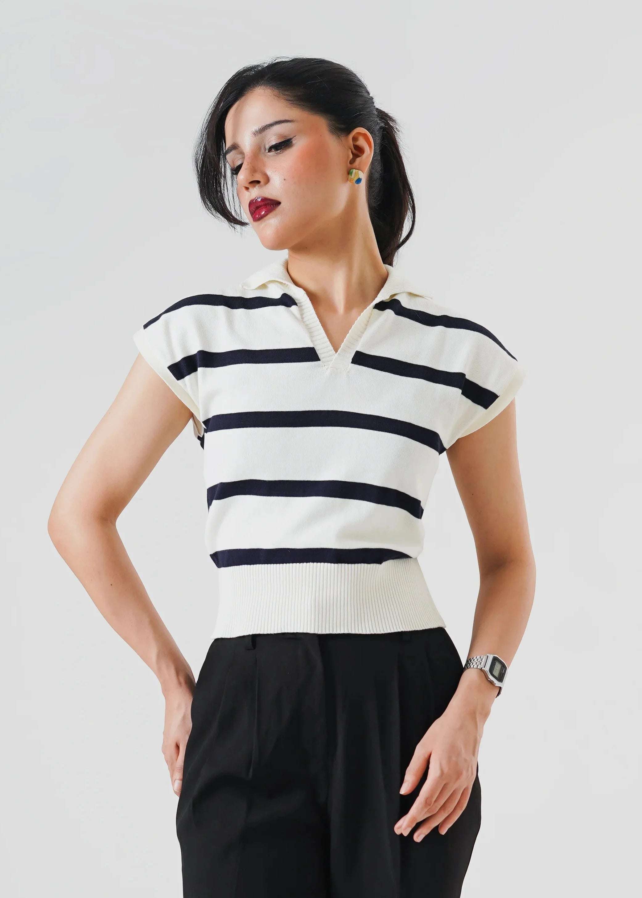 A woman modeling a short-sleeve polo shirt with horizontal black and off-white stripes, paired with black trousers and black heels, against a light gray studio background.