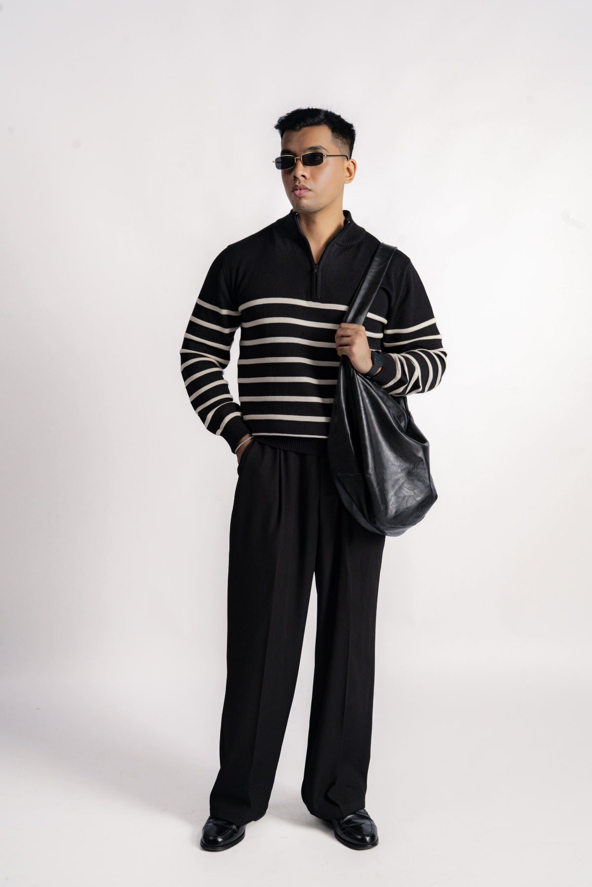 Man wearing a black and beige striped sweater with wide-leg pants, holding a black bag on a white background