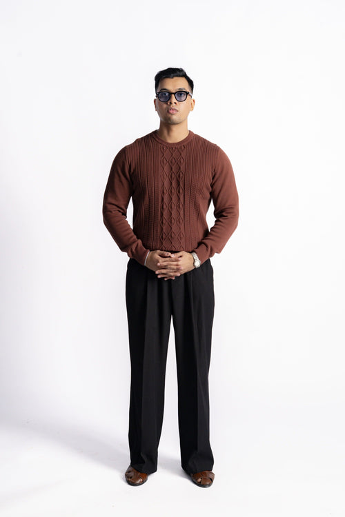 A man wearing a brown aran knit sweater with a cable pattern down the front, paired with black trousers and brown shoes, standing against a white studio background.
