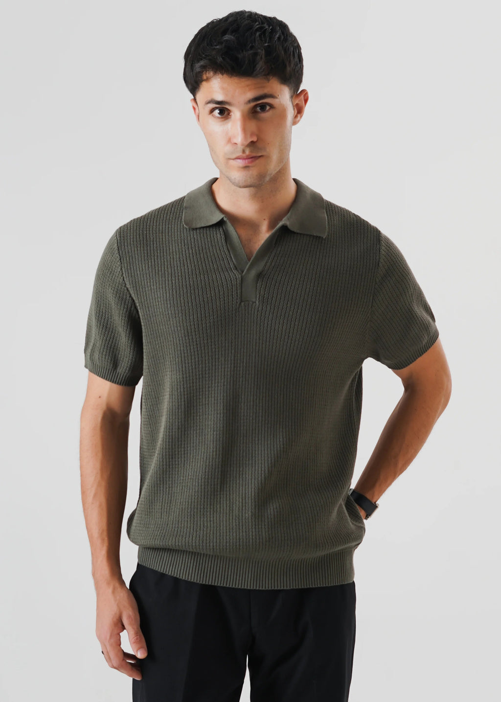 A man wearing a short-sleeve ribbed/racking knit polo shirt in olive green/khaki green, paired with black trousers, photographed against a light gray studio background.