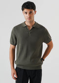 A man wearing a short-sleeve ribbed/racking knit polo shirt in olive green/khaki green, paired with black trousers, photographed against a light gray studio background.