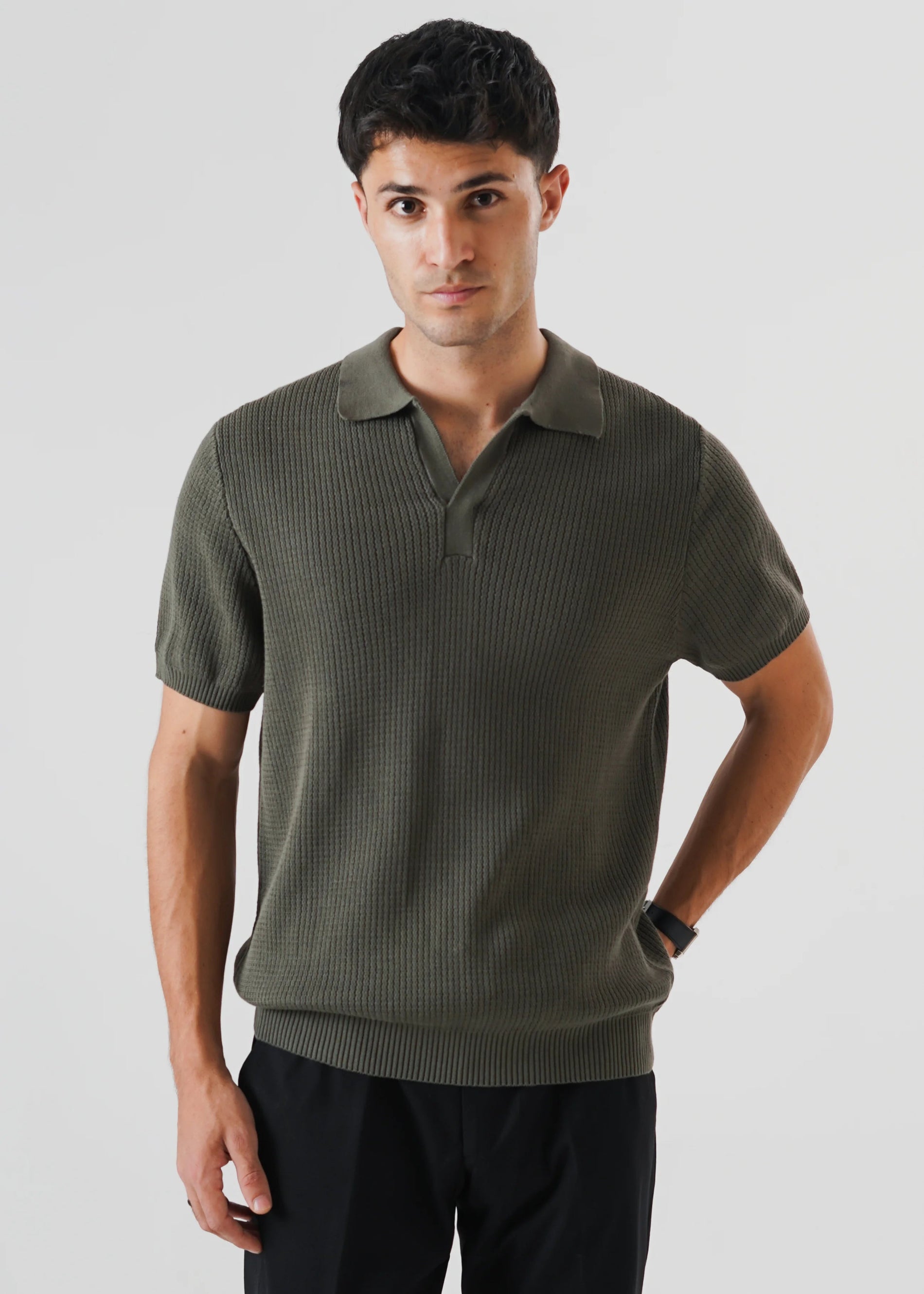 A man wearing a short-sleeve ribbed/racking knit polo shirt in olive green/khaki green, paired with black trousers, photographed against a light gray studio background.